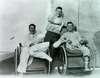 Three smiling veterans. Two leg amputees are seated in wicker wheelchairs. An arm amputee stands behind them.