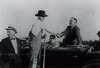 President Roosevelt sits up on the back of a car seat and shakes hands with a farmer in overalls standing next to the car.