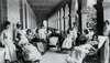 Seven women sit in large wheelchairs on the porch of a large Victorian house at a sanitarium. Each is covered in a blanket and looks passively at the camera. Six attendants and two children stand behind and amongst them.