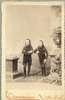 Two small-statured men with long hair and beards stand between two pedestals.