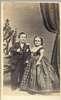A short-statuer man and woman stand between a chair and drapery.