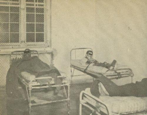 An Image of two beds. In each bed is a male presenting person who has straps holding them down across the waist with further restraints binding the feet together and to the bed, as well as the hands.