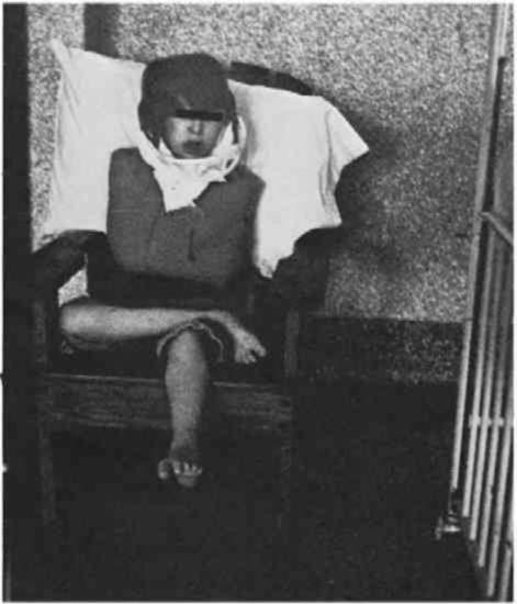 A Young person with dark hair is tied to a wooden chair, a restraining jacket additionally used to restrain them.