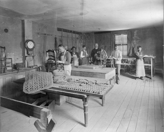 Workshop for the Blind, Perkins Institution for the Blind, Fourth Street, South Boston, Mass. In front are two thick door mats and a spool of the thick cord used to make them. Six men stand in the back of the room at work stations. Some work on chair caning. One of the faces has been enhanced with pen for publication purposes.