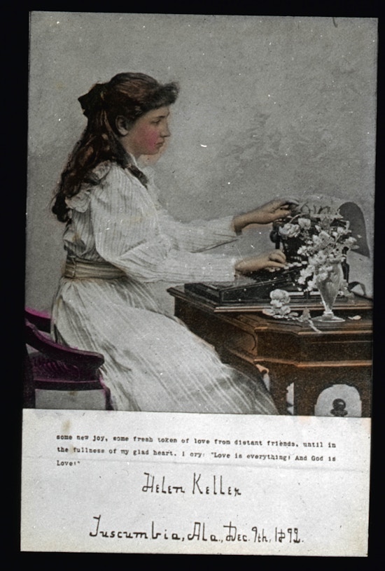 Young Helen Keller seated facing right, wearing a long sleeved dress at a table typing on a typewriter or braille writer.  In color.