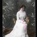 Helen Keller seated wearing a formal white gown with pink ribbon around the waist and holding flowers.