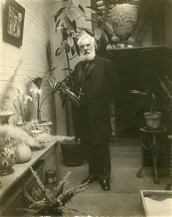 Alexander Graham Bell, standing indoors, looking at camera, surrounded by potted plants.