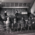 Photograph of Sarah Fuller's class with seated students facing chalkboard, away from camera. One student is standing at the board with Fuller instructing.