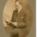 John Wright seated, looking down and left at possibly a manuscript or newspaper.
