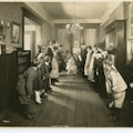 Students and teachers lining two sides of a hallway, looking away from camera, with a female wearing a white dress crouched at the far end of the hallway holding a round object at the center of attention.