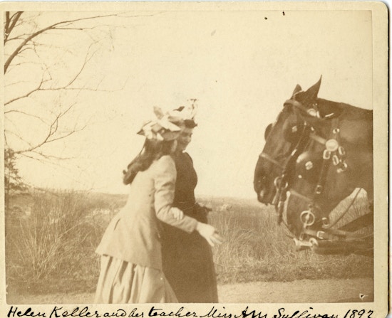 Helen Keller and Anne Sullivan (both left) stand together facing horses positioned right in Boston suburbs.