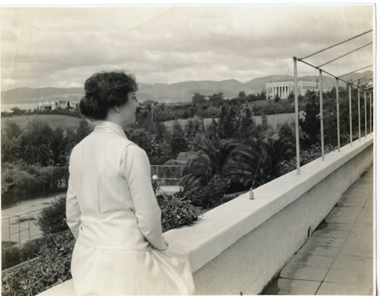 Helen Keller in a long sleeved, light colored dress standing outside, overlooking an expansive view of trees and hills in background.