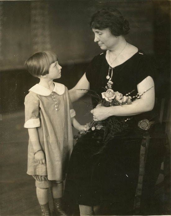 Helen Keller (right), seated, holding a bouquet of flowers, with child, standing.