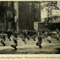 Men doing calisthenics in a field next to a three-story brick building.