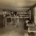 Boys' caning shop, Main building, Perkins Institution, South Boston, Mass. Showing Mr. Thomas J. Carroll, instructor. The boys stand at tall work benches with chair seats to be caned clamped at chest height. They each have long lengths of caning materials that they weave across the wooden frames.