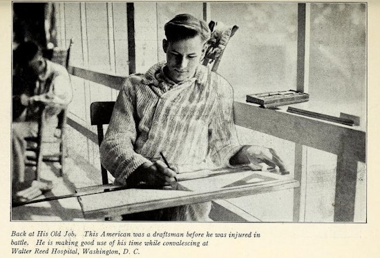 Young man sits at drafting table next to large window.