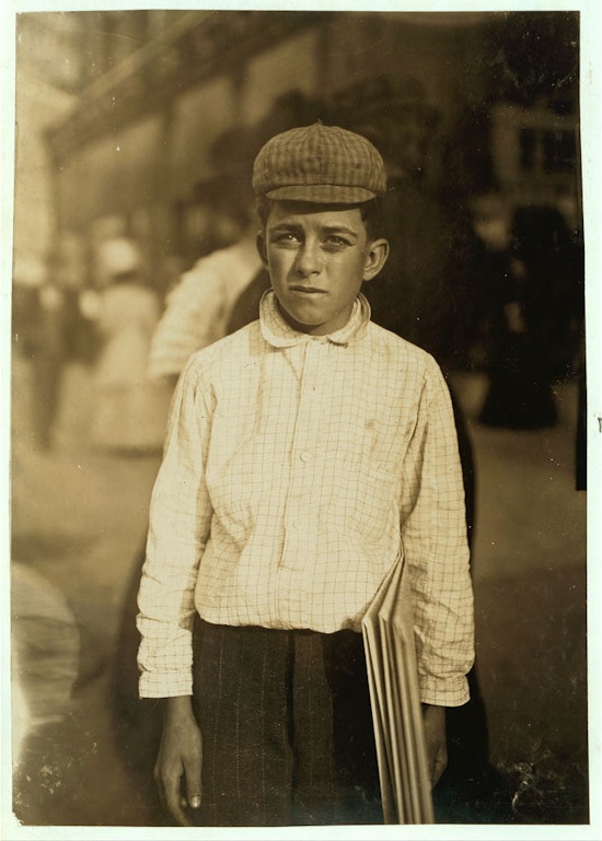 Boy with cap stands in street holding a newspaper.