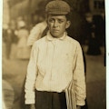 Boy with cap stands in street holding a newspaper.