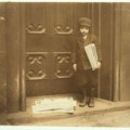 Young boy in hat stands before large door holding newspapers.