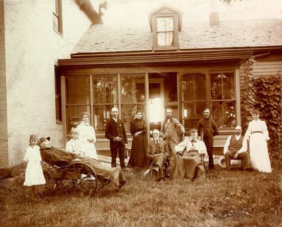 A family poses in front of the porch of a house.  An elderly man lies in a wheel chair.