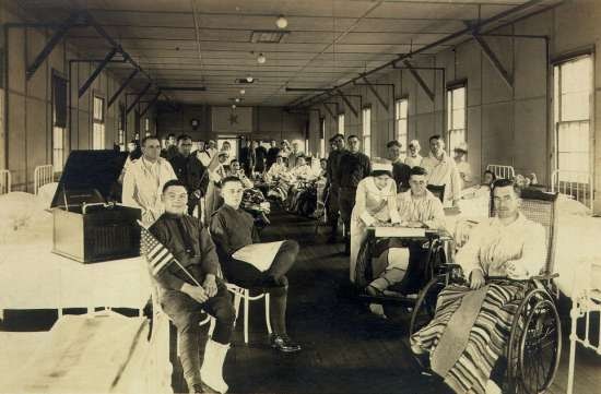 Photograph of ward in military hospital, including veterans, nurses, and doctors.  Several men use wheelchairs.
