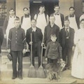 Photograph of men, one woman, one child, and a dog posing on the porch of a building.