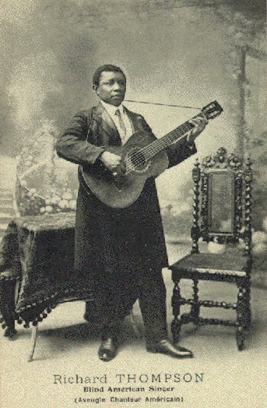 A black man wearing long coat and necktie stands playing a guitar in front of a table and a chair