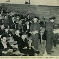 A group of civilian men sit on benches as two men in military uniforms hand out exams