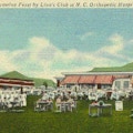 Colored postcard of staff and patients eating watermelon on lawn in front of low buildings with red and white awnings