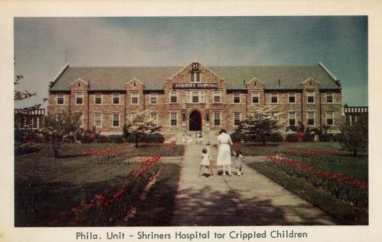 A woman leads two little girls, one who uses crutches, up the front walk past flowers to the hospital.