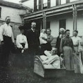 Photograph of a family posing outside of a house.  A young man wearing a night shirt sits in a wooden box.
