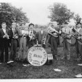 A nine-piece brass band stands in a field in front of trees holding their instruments.  They stand around a large drum on which is printed State Insane Asylum Band, Topeka, KS.