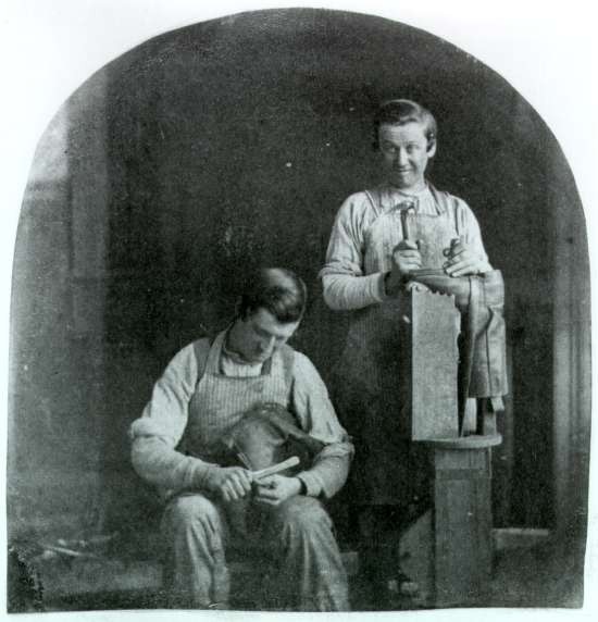 Two boys working as cobblers. One files the sole of a shoe. The other holds a hammer and last upright and smiles.
