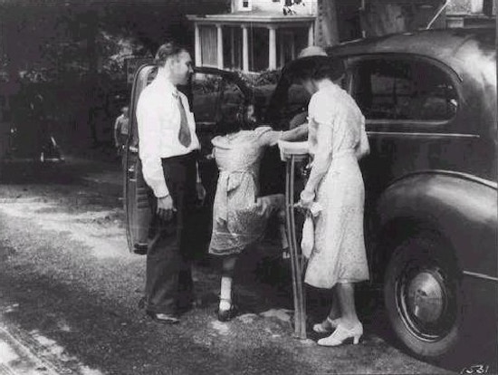Little girl with braces on her legs climbs into car; man stands to her left and a woman, holding crutches, stands to her right
