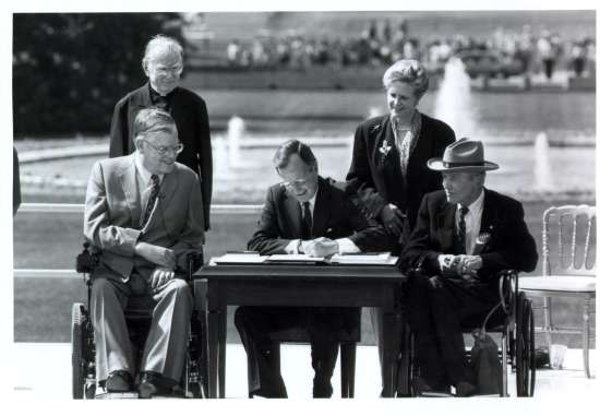 George Bush in front of White House sitting at table signing ADA; with a priest, a woman, and two men in wheel chairs