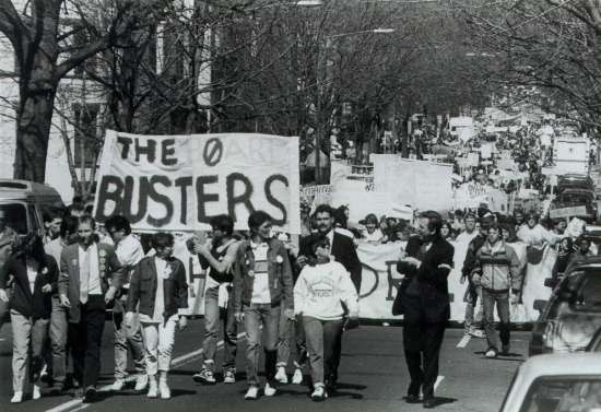 A crowd of protesters marches down a street.  A man holds a banner that reads "The Board Busters."