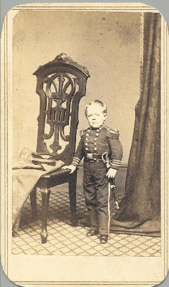 A short-statured young man in a military uniform stands next to a chair.
