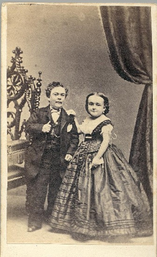A short-statuer man and woman stand between a chair and drapery.