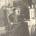 A man in a wooden wheelchair looks up from his desk.
