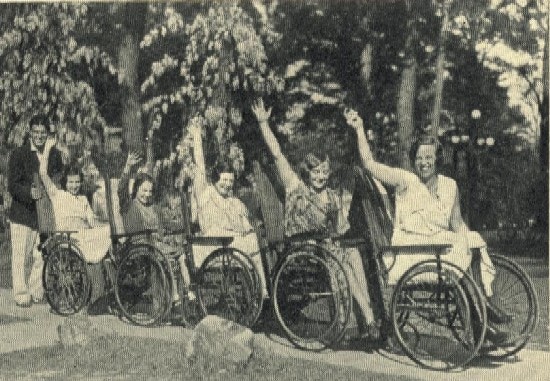 Five women in a row of wheelchairs, the last pushed by a young man.