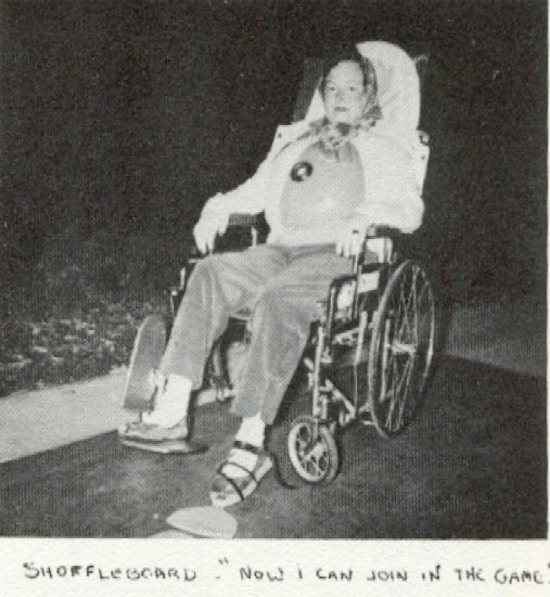 A woman in wheelchair and wearing a chestliece plays shuffleboard.