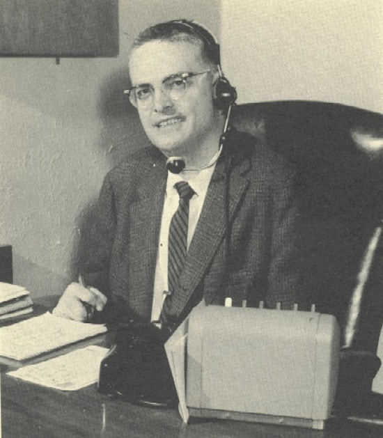 A man in a suit sits at a desk and wears a telephone headset.