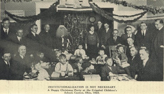 A group of children with presents, surrounded by adults, one dressed as Santa Claus.