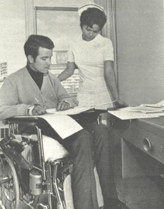 A man in a wheelchair and a nurse look at papers.