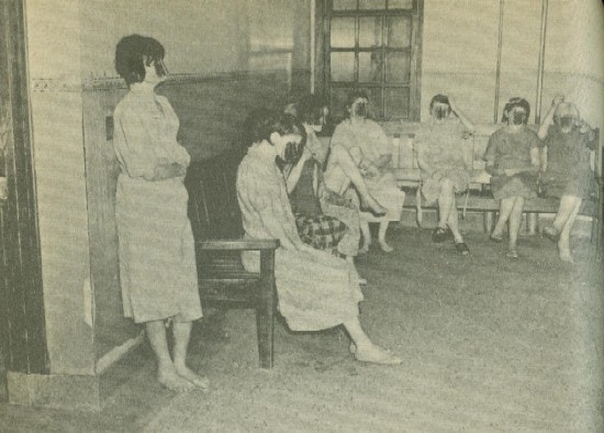 Seven women, most barefoot, sit on benches in a room.