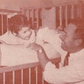 A smiling father plays with a young girl in a crib.
