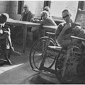 Men in a ward, some around a table, one in a wheelchair.