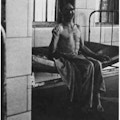 A partially clothed man sitting on a bed next to a barred window.