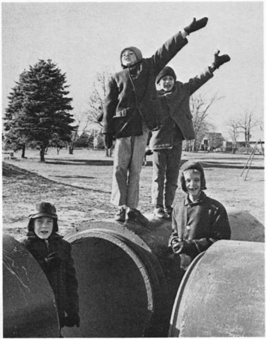 Three boys near large pipes, two saluting.