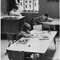 A teacher at her desk and two children at tables.